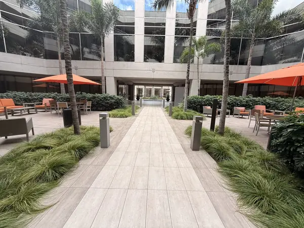 A tiled walkway leading to the building lobby, between planted borders, palm trees and tables shaded by orange umbrellas.